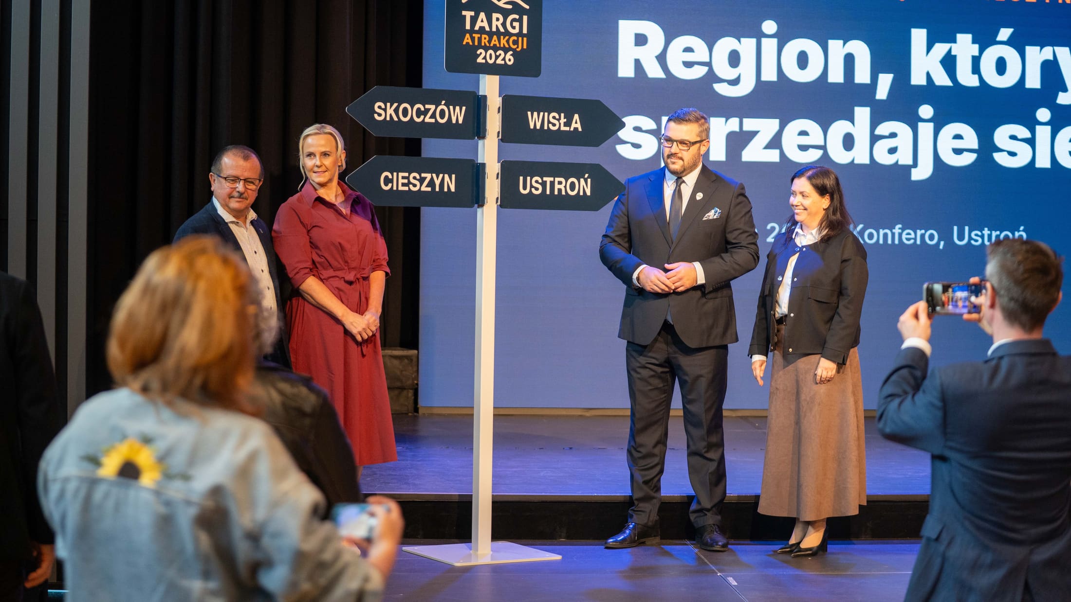 Przedstawiciele miast patronów honorowych Targów Atrakcji stoją wspólnie przy scenicznym drogowskazie z logo wydarzenia i nazwami Skoczów, Ustroń, Cieszyn i Wisła